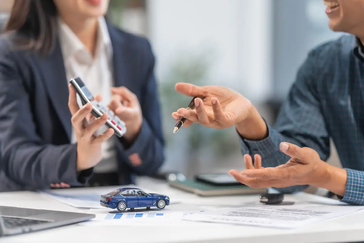 Zwei Personen sitzen am Schreibtisch und besprechen einen Autokauf oder eine Finanzierung. Eine hält einen Taschenrechner, die andere einen Stift; auf dem Tisch liegen Unterlagen, ein Laptop, Autoschlüssel und ein kleines blaues Modellauto.