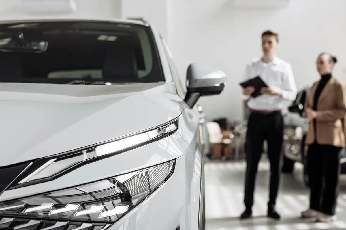 Nahaufnahme der Front eines weißen SUVs im Autohaus. Im Hintergrund stehen unscharf zwei Personen, eine hält ein Tablet und bespricht offenbar den Autokauf.