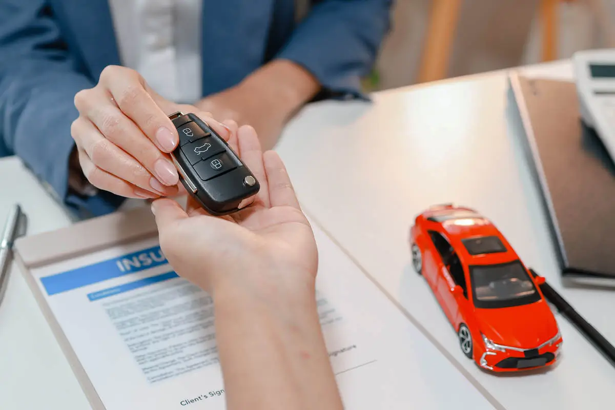 Schlüsselübergabe am Schreibtisch: Eine Person reicht einem anderen den Autoschlüssel über Vertrags- bzw. Versicherungsunterlagen. Daneben liegen ein rotes Modellauto, ein Stift und ein Taschenrechner.