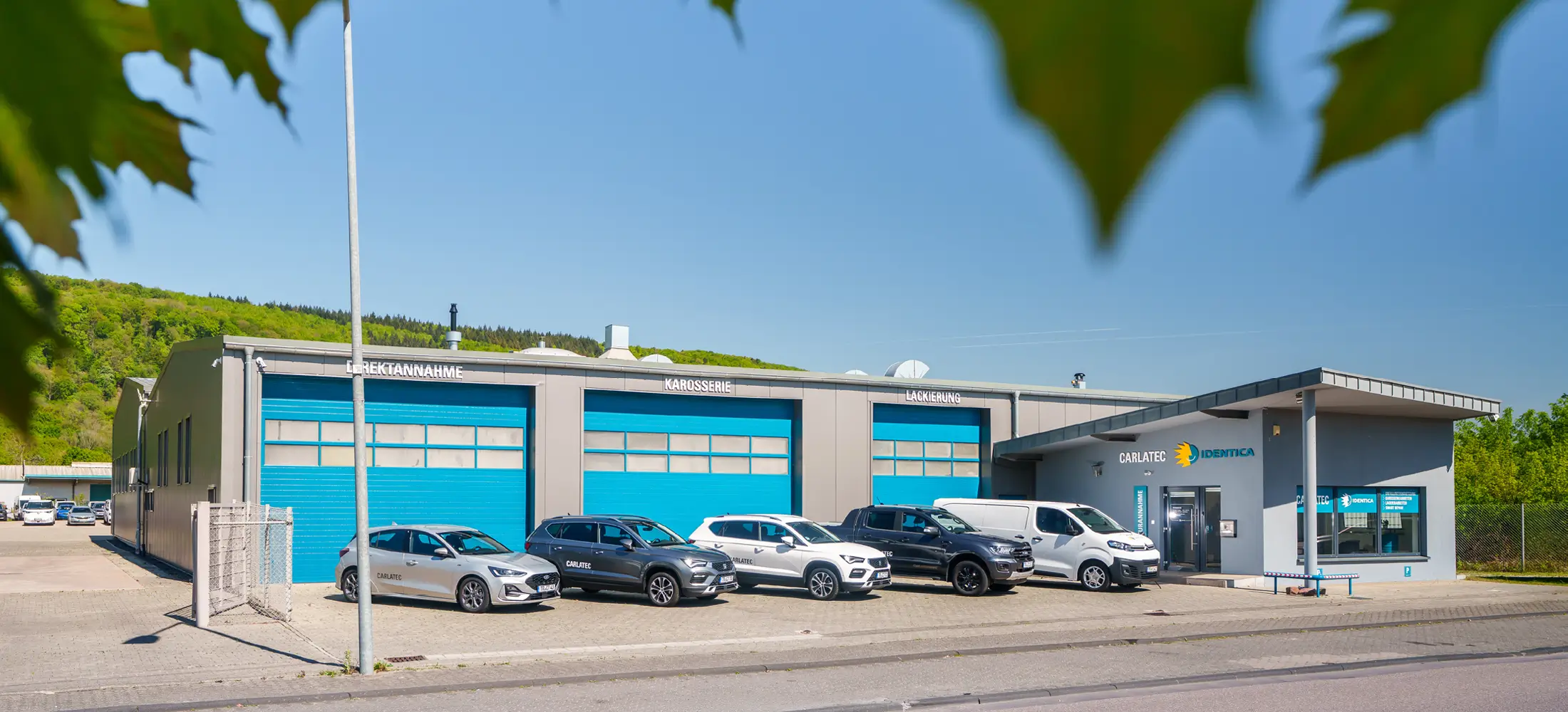 Gewerbegebäude einer Kfz-Werkstatt mit blauen Rolltoren und den Beschriftungen „Direktannahme“, „Karosserie“ und „Lackierung“. Mehrere Autos und Transporter parken davor.
