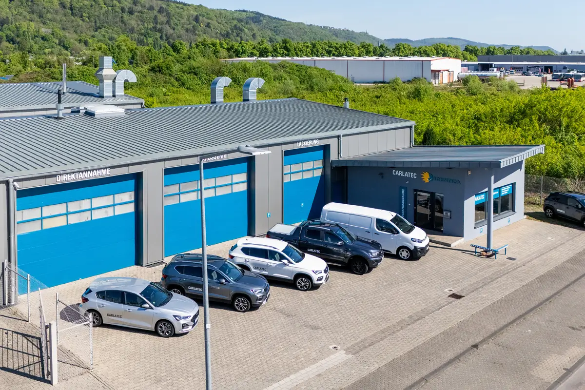 Gewerbegebäude einer Kfz-Werkstatt mit blauen Rolltoren und den Beschriftungen „Direktannahme“, „Karosserie“ und „Lackierung“. Mehrere Autos und Transporter parken davor.