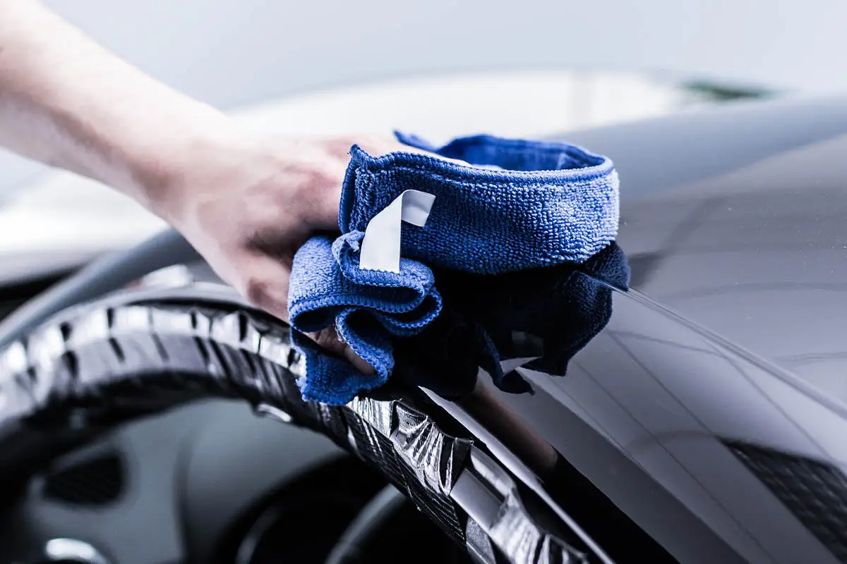 Eine Hand poliert die lackierte Oberfläche eines Autos mit einem blauen Mikrofasertuch; die Kanten sind mit Klebeband abgeklebt.