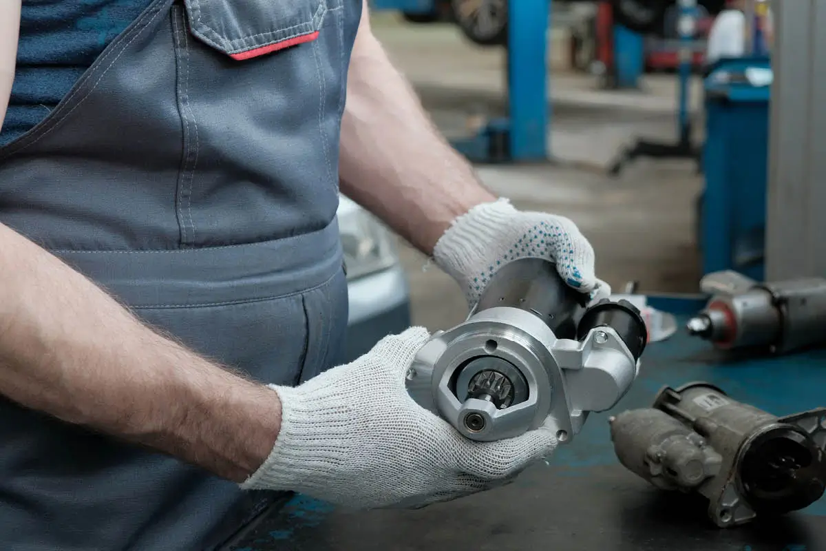 Ein Mechaniker in der Werkstatt hält mit weißen Arbeitshandschuhen einen Anlasser in den Händen. Auf der Werkbank liegen weitere aus- oder einzubauende Teile, der Hintergrund ist unscharf.