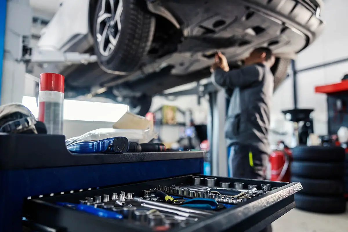 In einer Autowerkstatt steht ein Wagen auf der Hebebühne. Im Vordergrund sieht man einen Werkstattwagen mit Steckschlüsseln und Werkzeugen, im Hintergrund arbeitet unscharf ein Mechaniker am Unterboden des Autos.